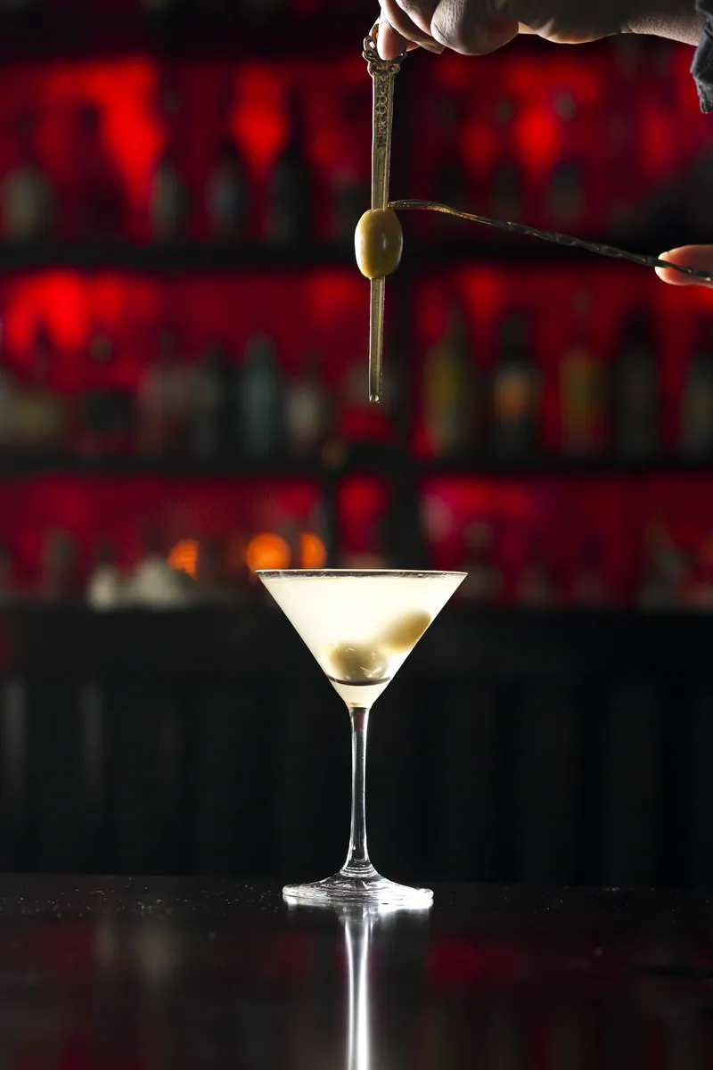 A martini with an olive dropping in, against a red-lit bar